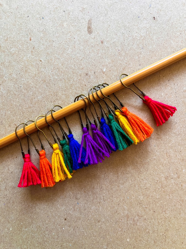 A rainbow of tassel stitch markers on a knitting needle.